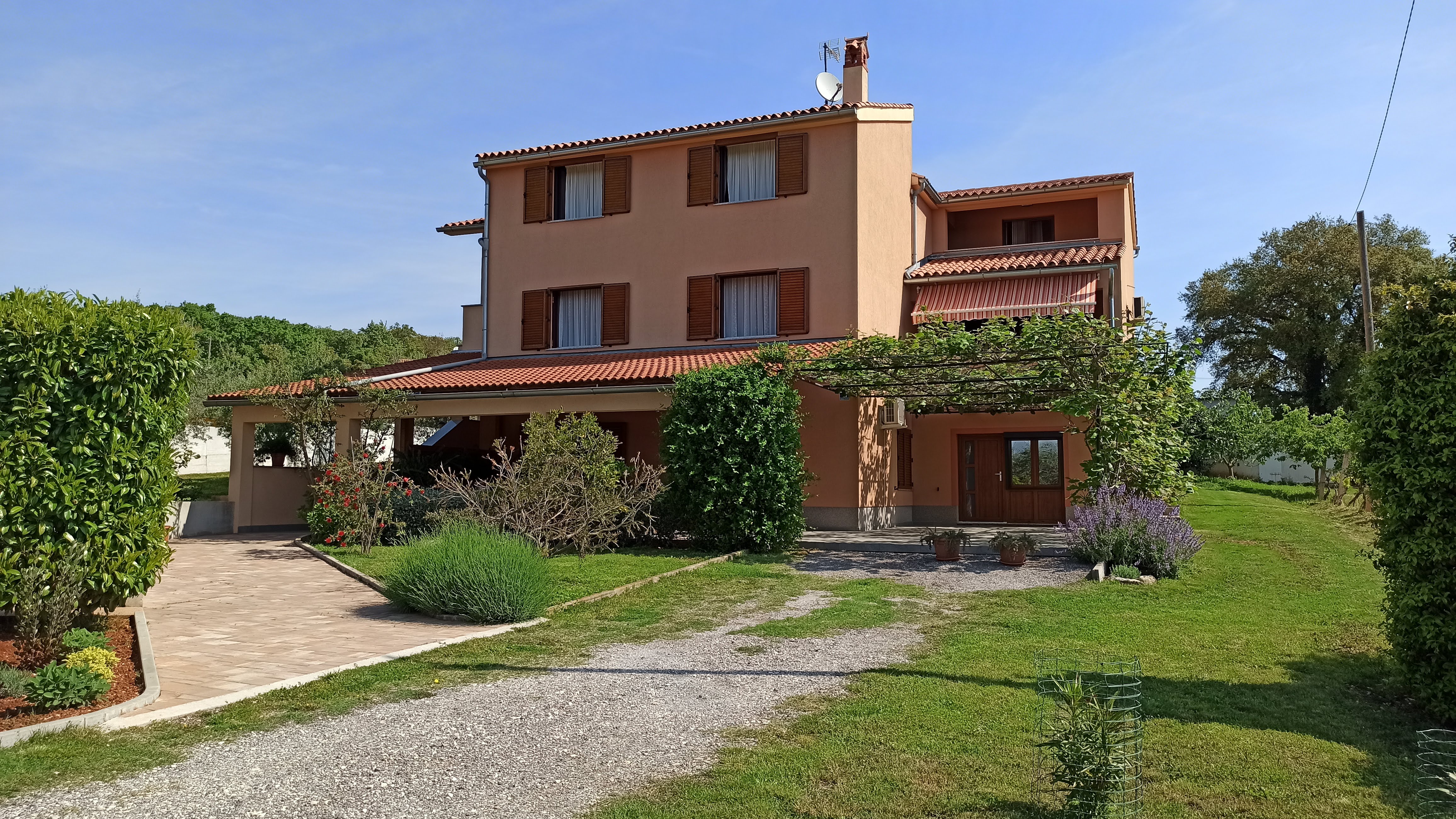 Beautiful colorful sunny garden, spacious parking space and a tiled access path. Colorful flowers filling the garden. A covered terrace and pergola in front of the apartment's entrance . Olive grove and nature in the background.