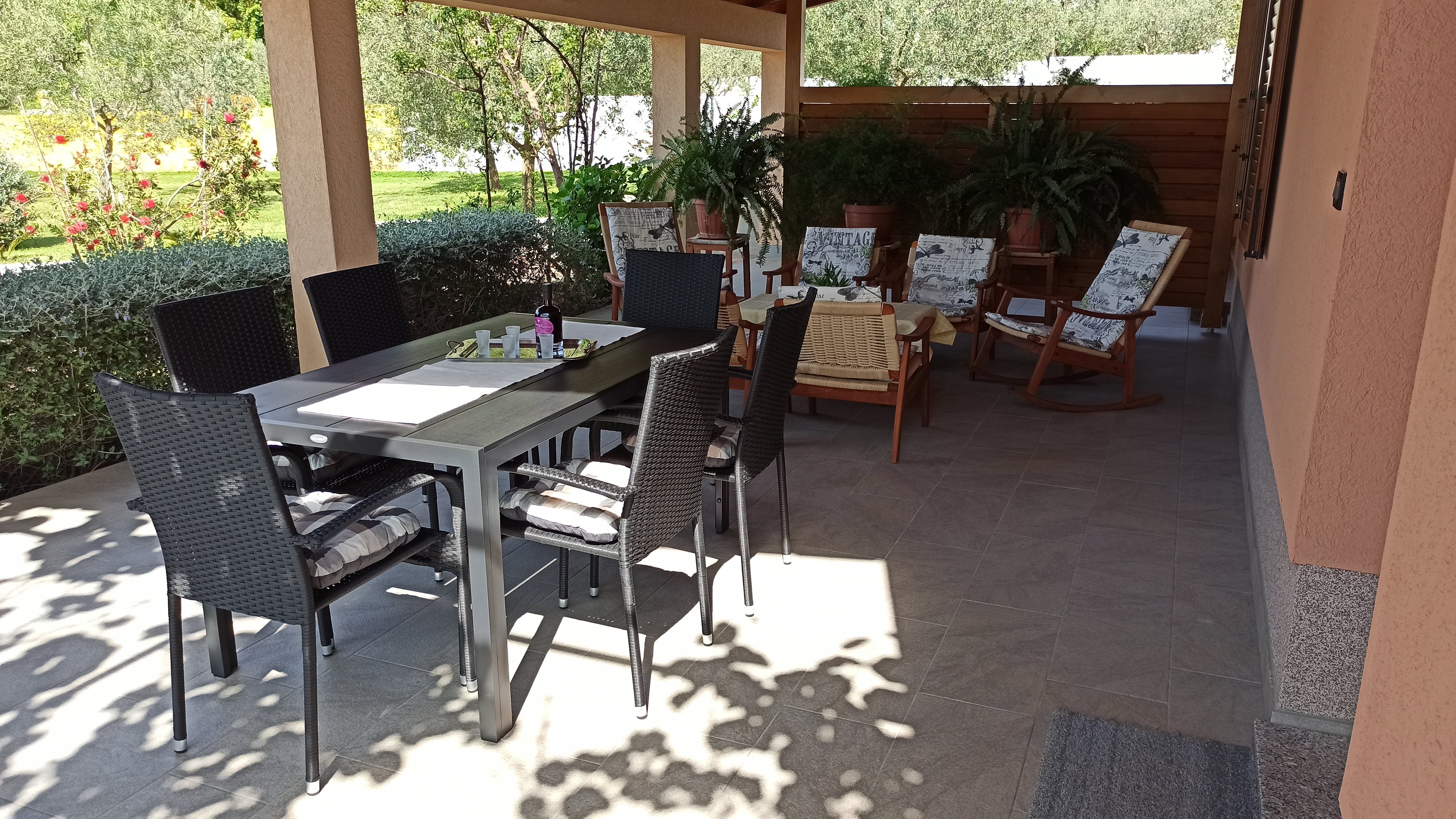 Bottle of wine and glasses on a tableclothed dining table. White platic chairs around the table. Bright covered terrace in the background. Armchairs covered with pillows. Large openeg window with curtains. Wooden barrier and green plants.