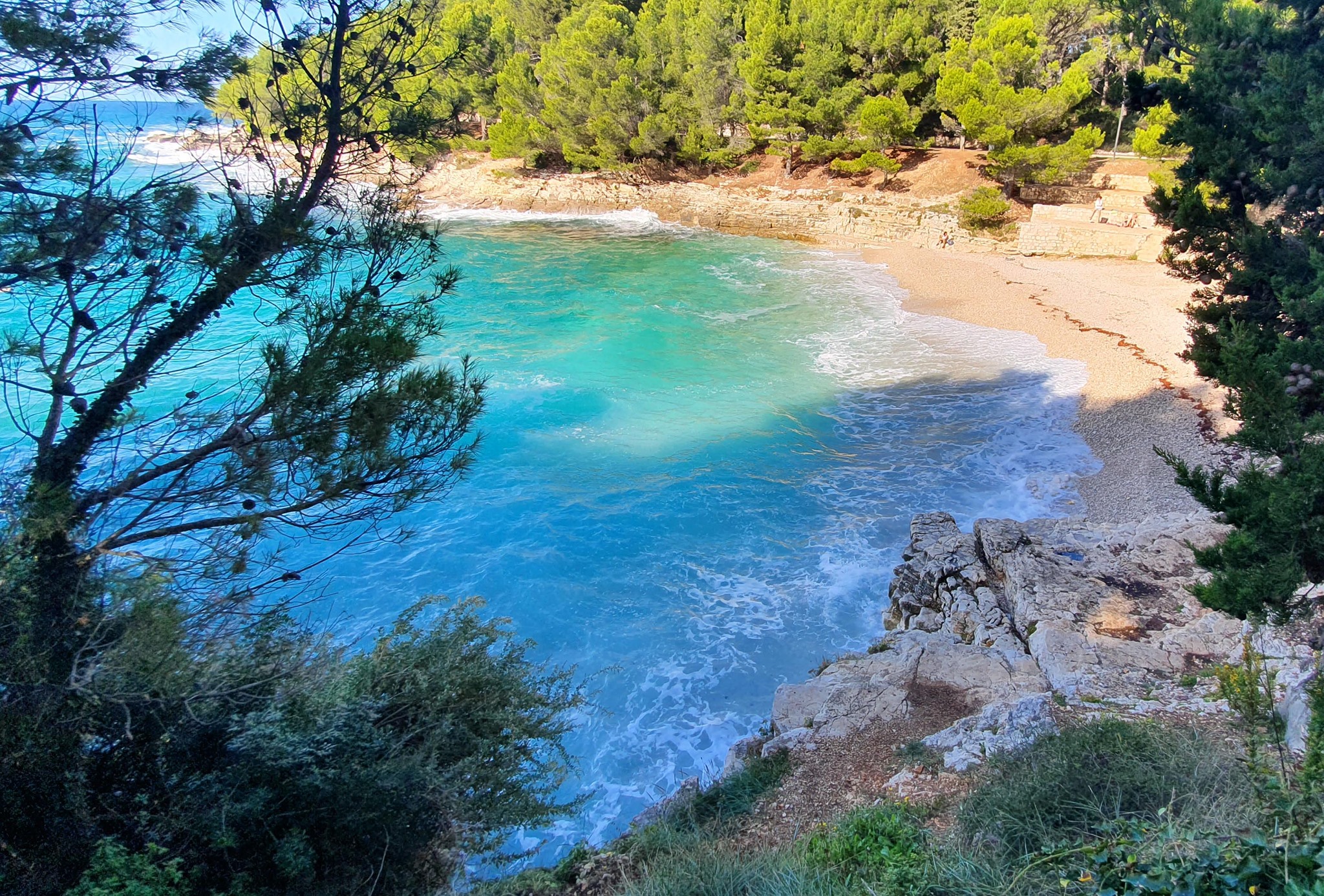 Gortanova beach in Pula near Apartments Borina. It is a pebbled beach with cliffs on each side, stairs and forest all around it. View from a cliff which has bushes on top. Clear, clean and blue sea with waves. Stairs and nearby road.