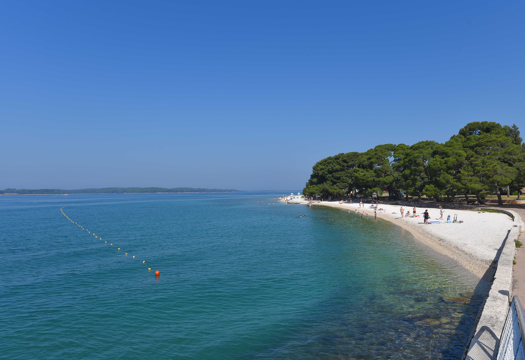 Long shoreline of the Valbandon beach in Valbandon near Apartments Borina. Pebbled beach. Blue sea and sky. View from a bridge. Clear and clean sea. People along the beach. Sea beacons along the shore. Forest and pathway in the background.